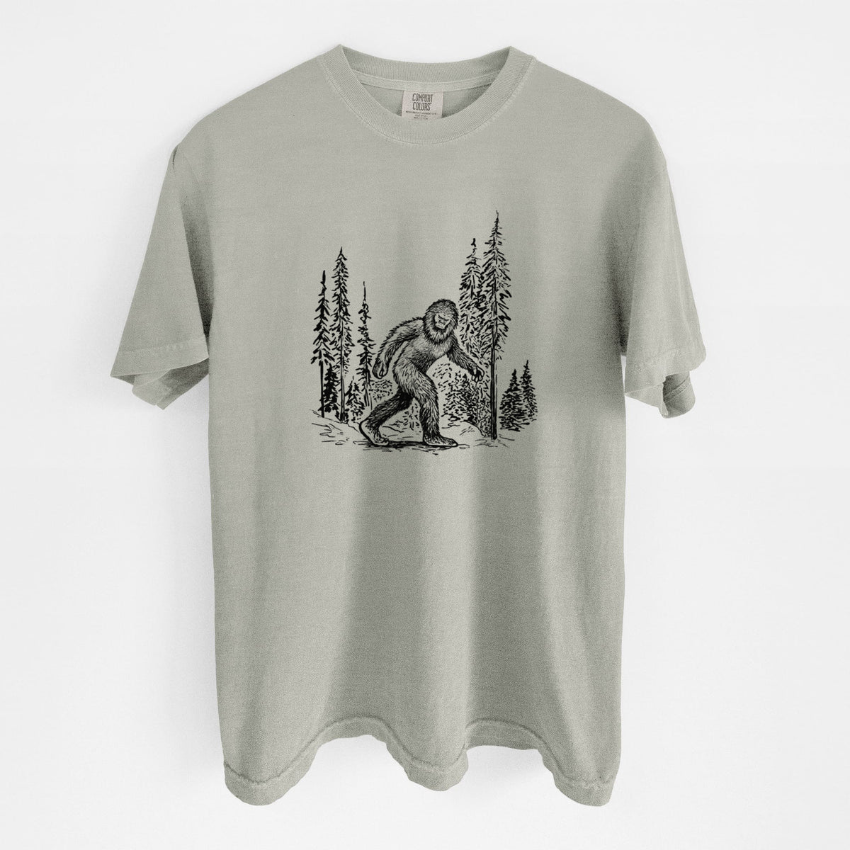 Sandstone beige Comfort Colors t-shirt laid flat, printed with a black ink illustration of Bigfoot walking through a pine forest.