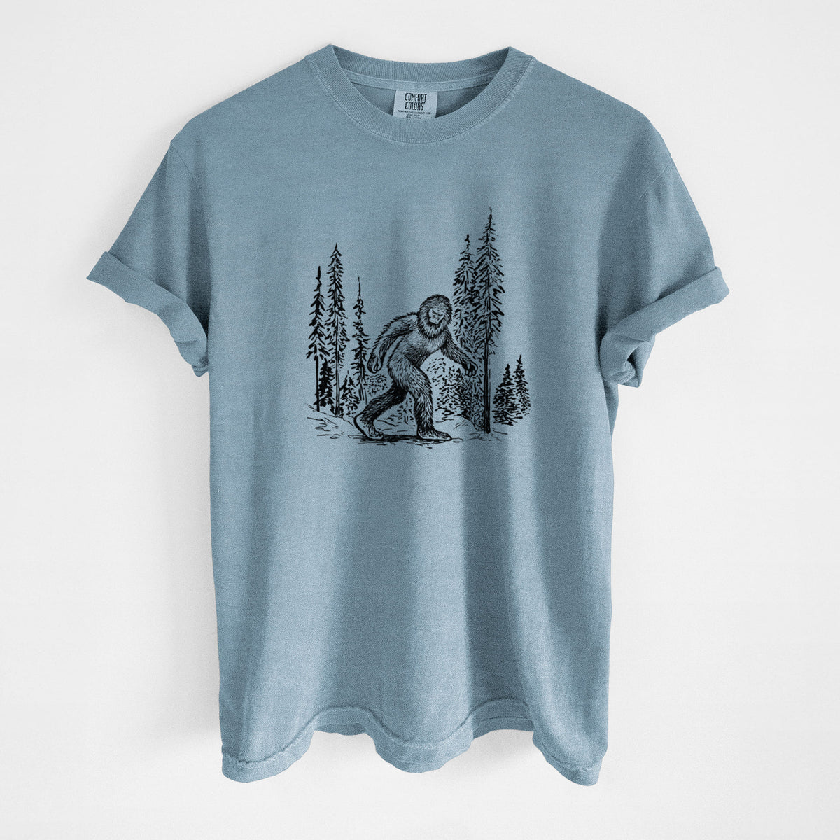 Steel blue Comfort Colors t-shirt laid flat, printed with a black ink illustration of Bigfoot walking through a pine forest.