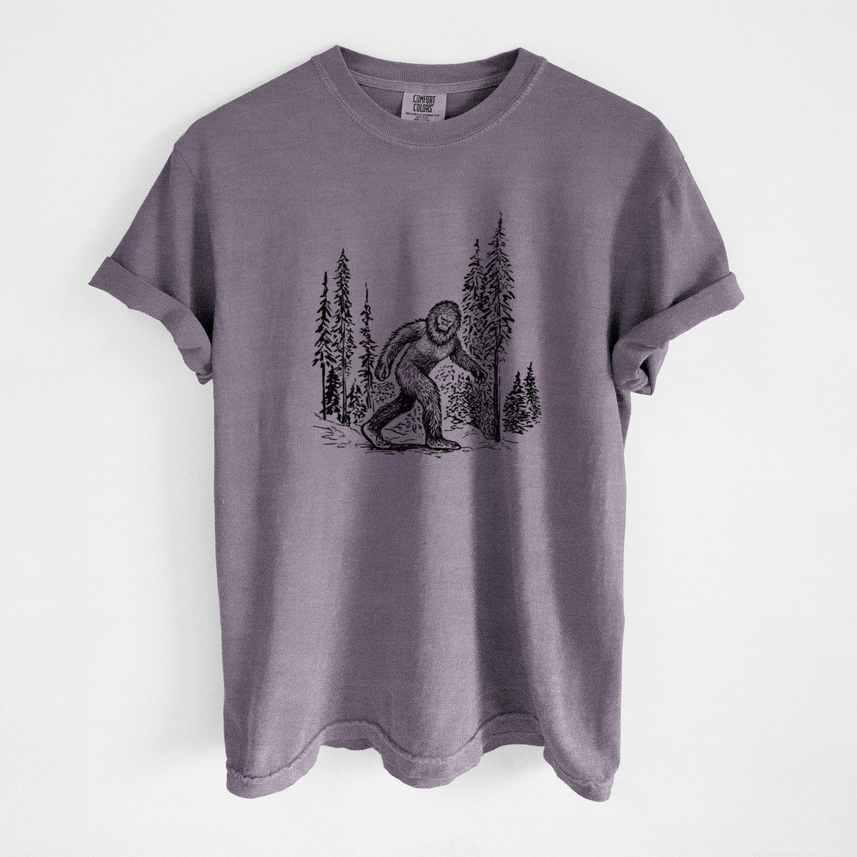 Wine red Comfort Colors t-shirt laid flat, printed with a black ink illustration of Bigfoot walking through a pine forest.