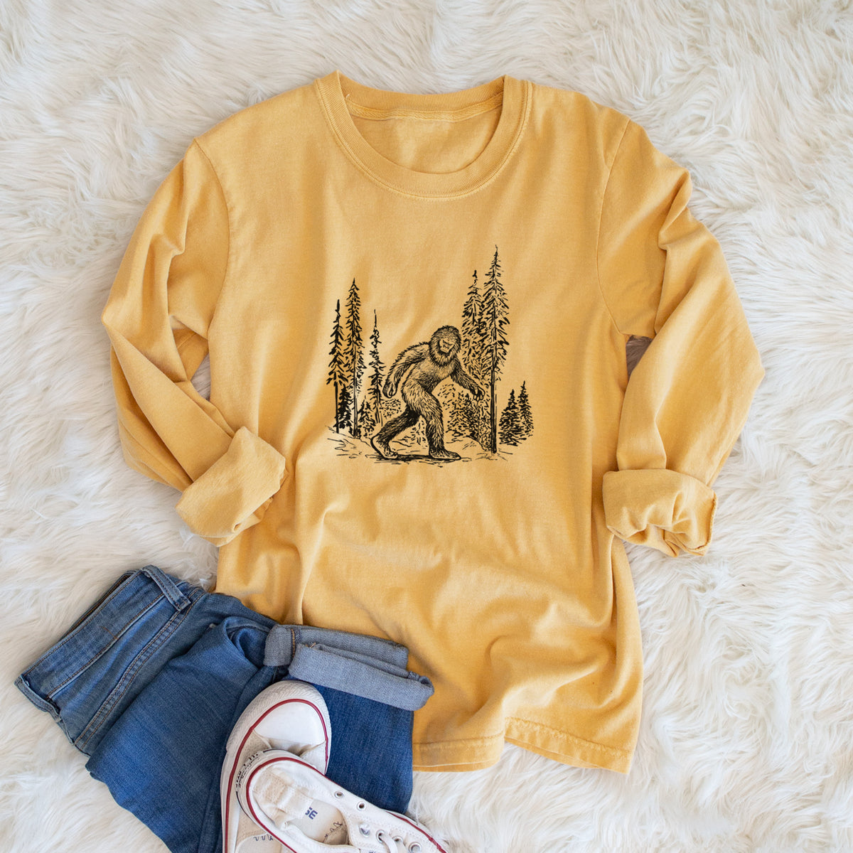 Golden wheat yellow Comfort Colors long sleeve shirt printed with a black ink illustration of Bigfoot walking through a pine forest, shown in a lifestyle photo.