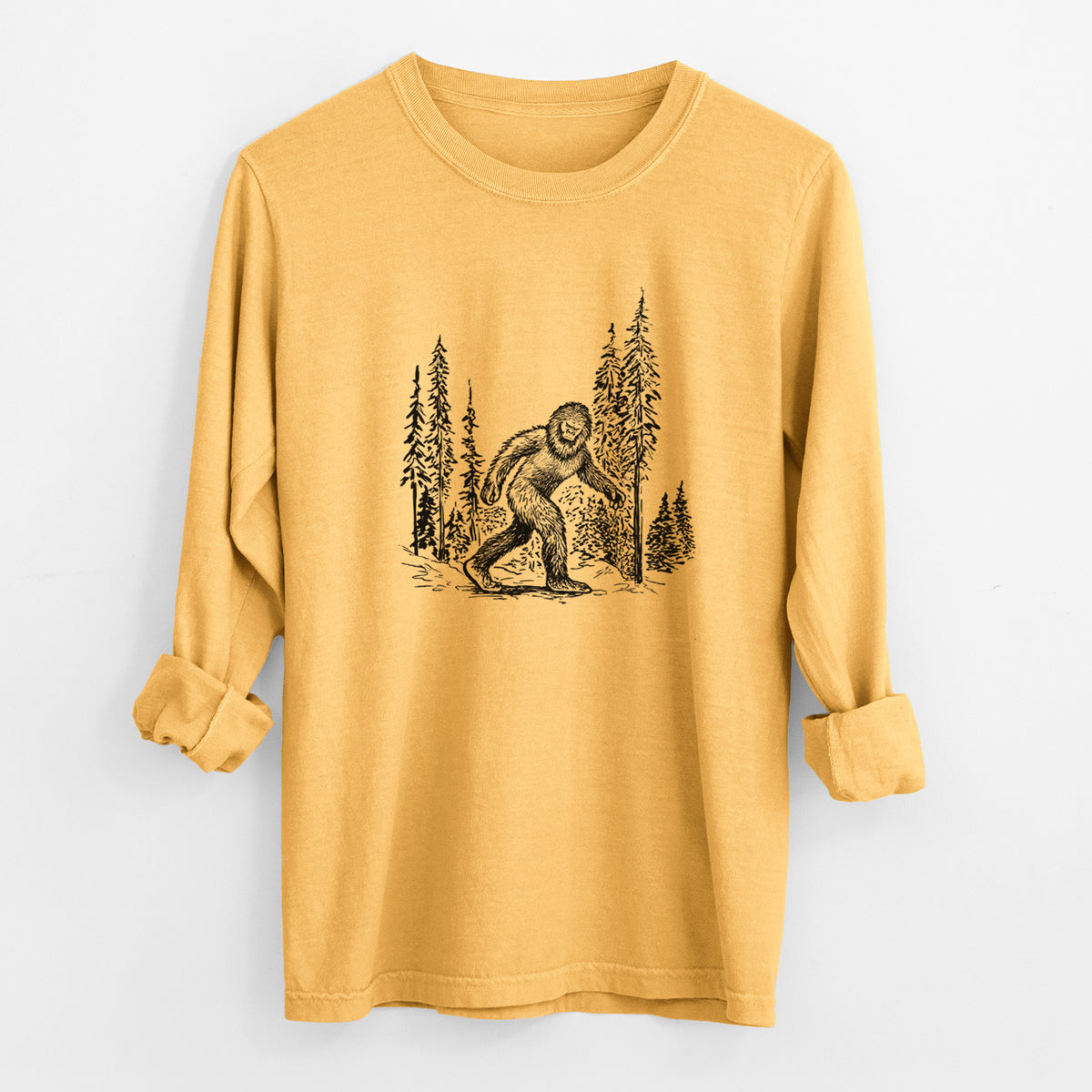 Golden wheat yellow Comfort Colors long sleeve shirt laid flat with the cuffs rolled, printed with a black ink illustration of Bigfoot walking through a pine forest.