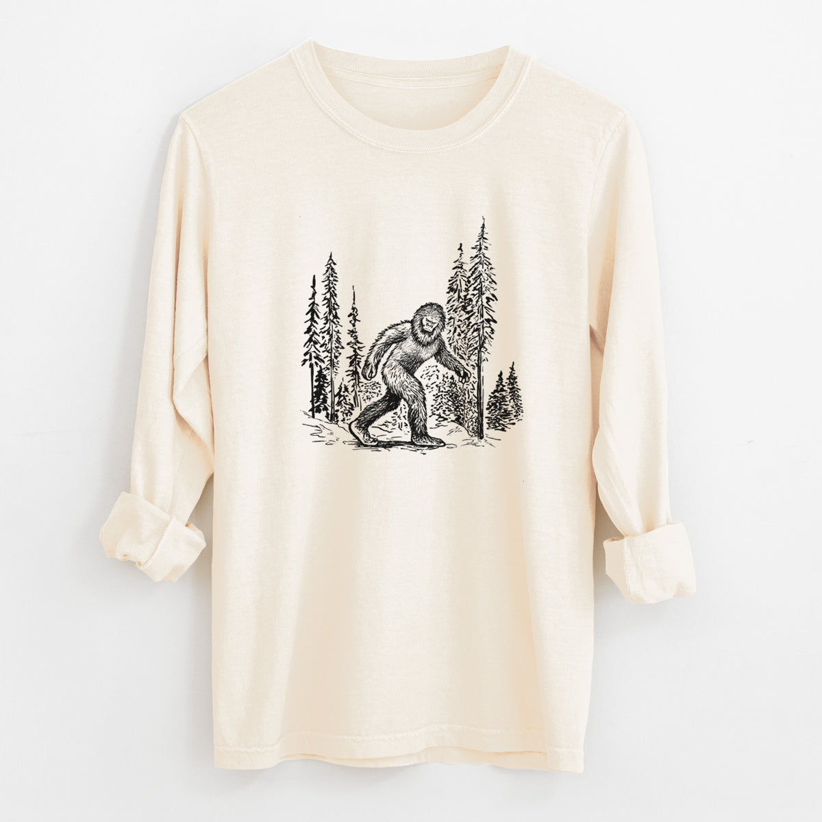 Ivory Comfort Colors long sleeve shirt laid flat with the cuffs rolled, printed with a black ink illustration of Bigfoot walking through a pine forest.