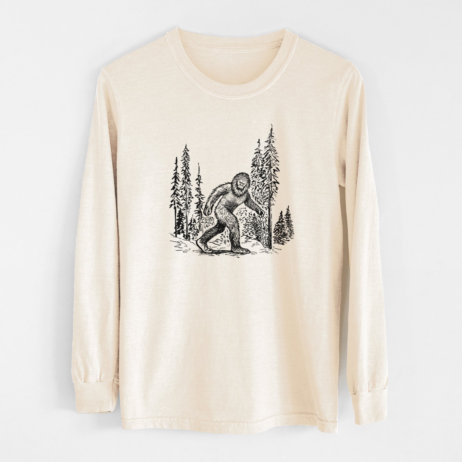 Ivory Comfort Colors long sleeve shirt laid flat, printed with a black ink illustration of Bigfoot walking through a pine forest.