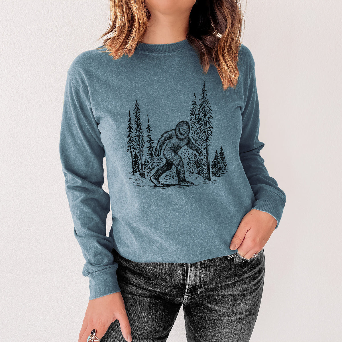 Steel blue Comfort Colors long sleeve shirt printed with a black ink illustration of Bigfoot walking through a pine forest, shown in a lifestyle photo.