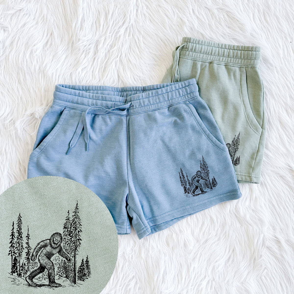 Two pairs of Cali Wave shorts in different colors printed with a black ink illustration of Bigfoot walking through a pine forest, with a close-up of the print.