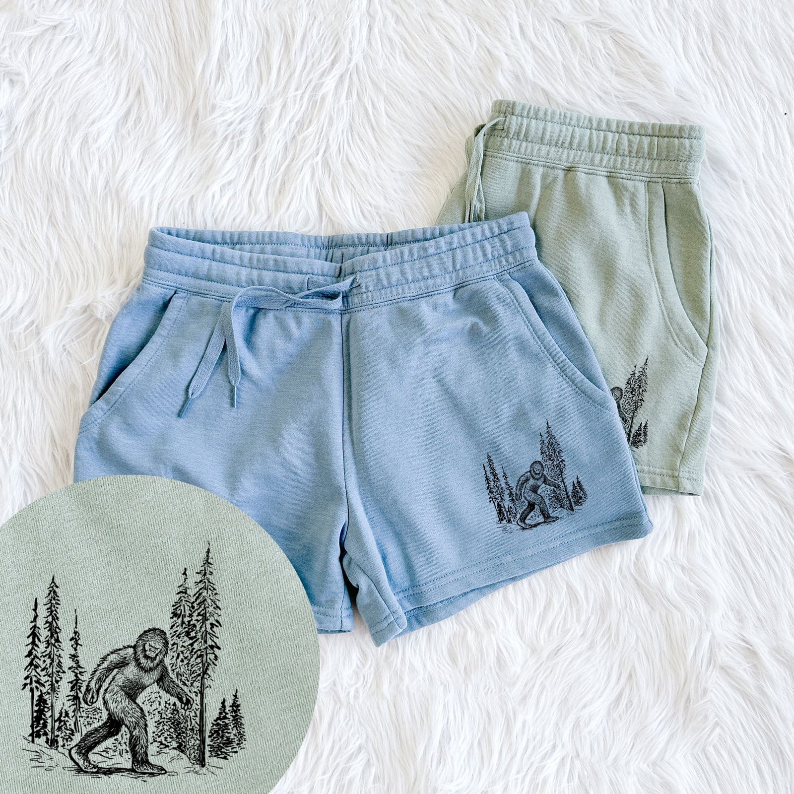 Two pairs of Cali Wave shorts in different colors printed with a black ink illustration of Bigfoot walking through a pine forest, with a close-up of the print.
