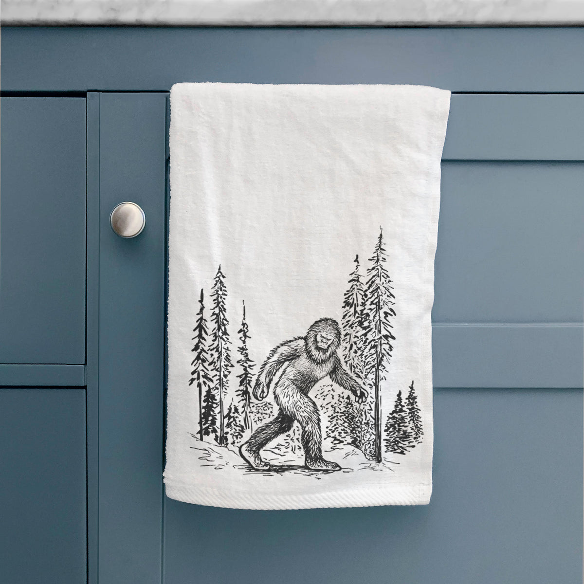 White hand towel printed with a black ink illustration of Bigfoot walking through a pine forest, hanging on a cabinet door.
