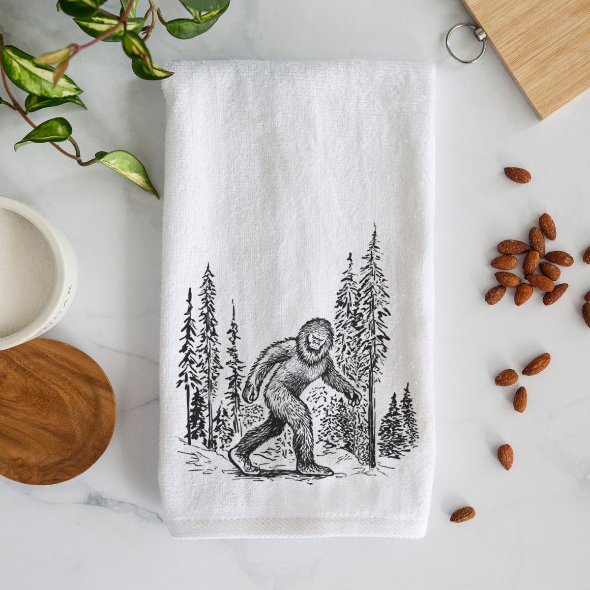 White hand towel printed with a black ink illustration of Bigfoot walking through a pine forest.