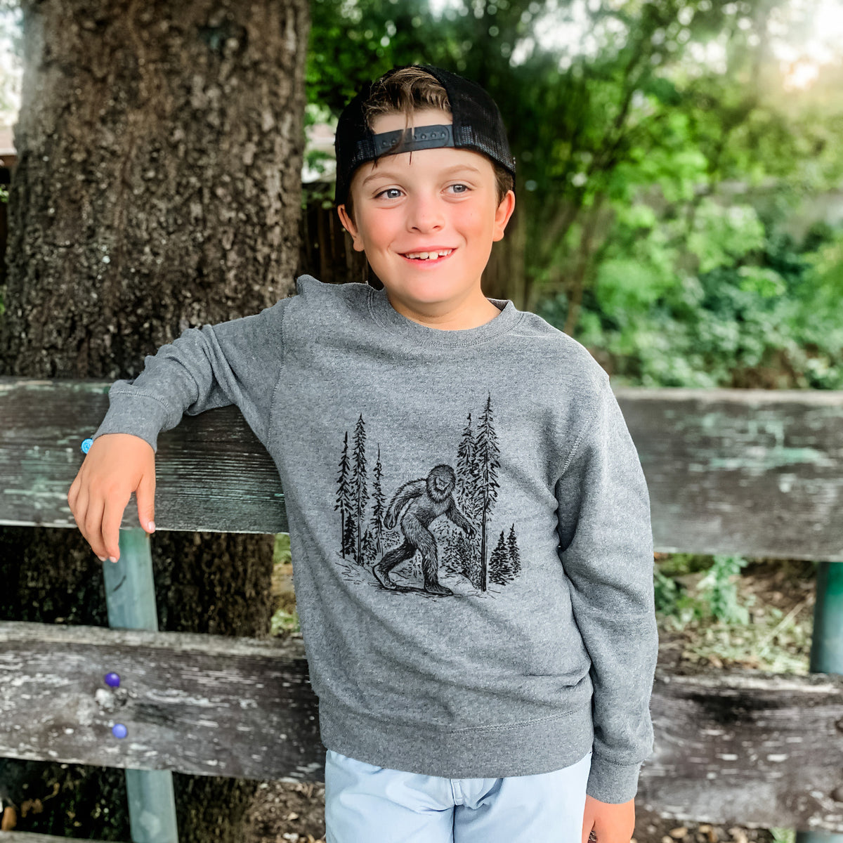 Boy wearing a youth crewneck sweatshirt printed with a black ink illustration of Bigfoot walking through a pine forest, outdoors.