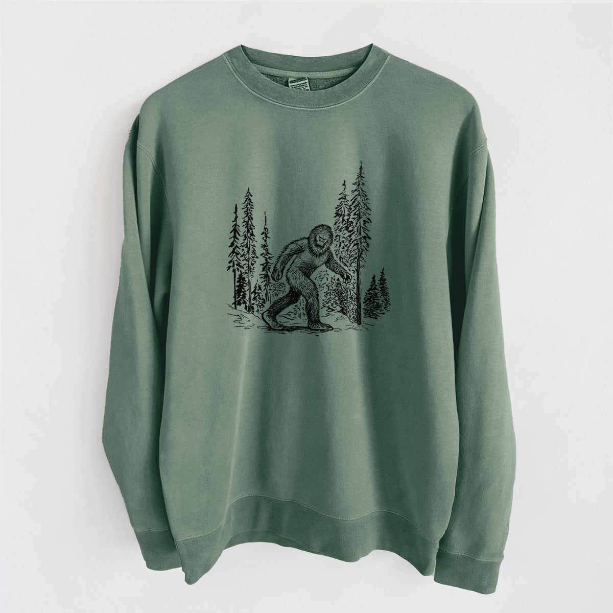 Alpine green pigment dyed crewneck sweatshirt laid flat, printed with a black ink illustration of Bigfoot walking through a pine forest.