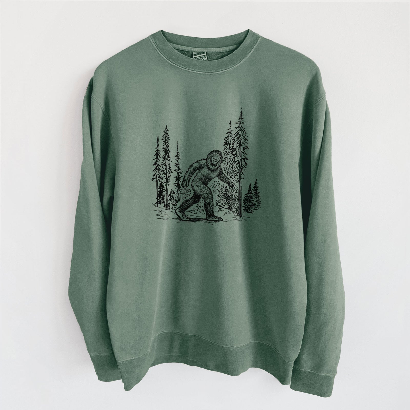 Alpine green pigment dyed crewneck sweatshirt laid flat, printed with a black ink illustration of Bigfoot walking through a pine forest.