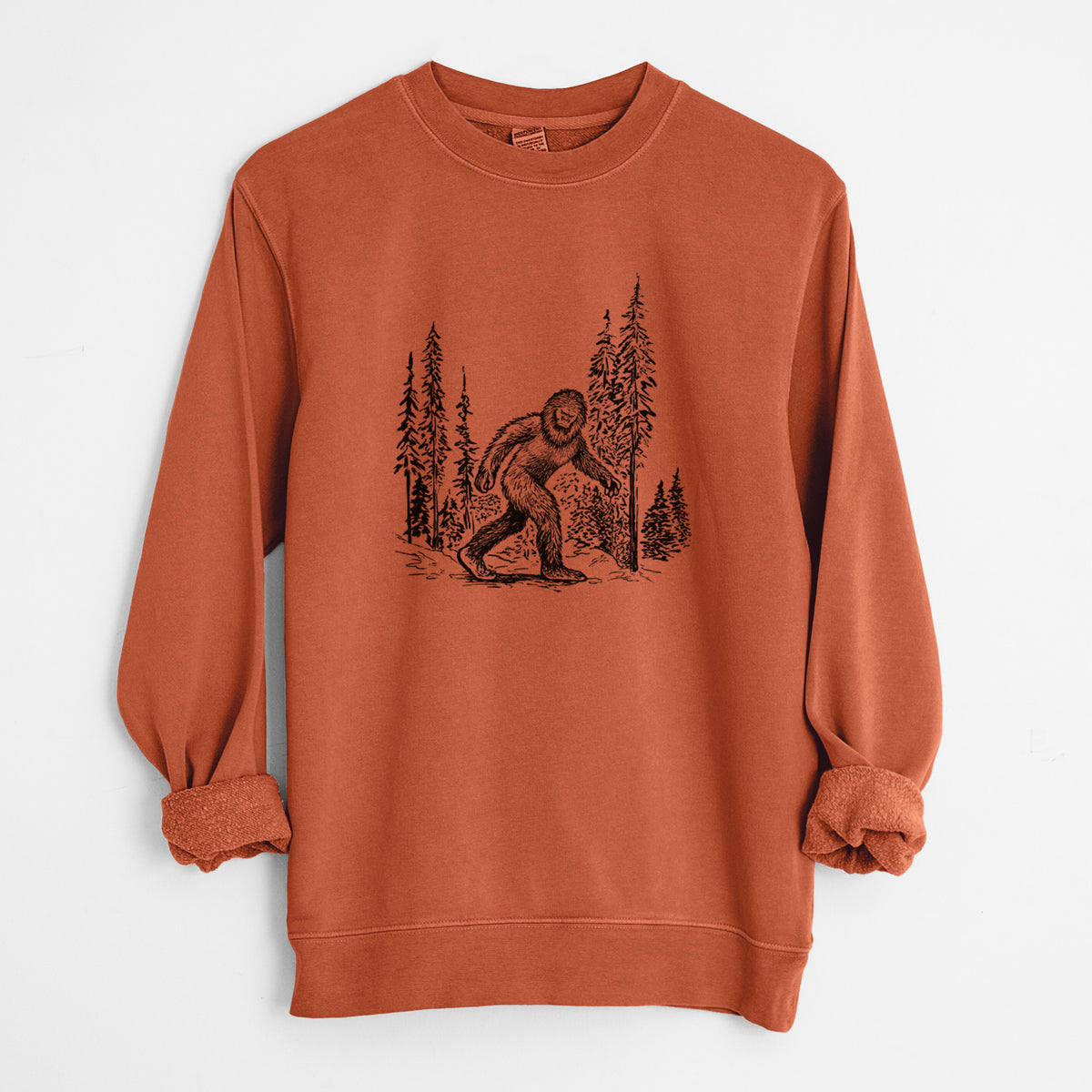 Amber orange pigment dyed crewneck sweatshirt laid flat with the sleeves rolled, printed with a black ink illustration of Bigfoot walking through a pine forest.