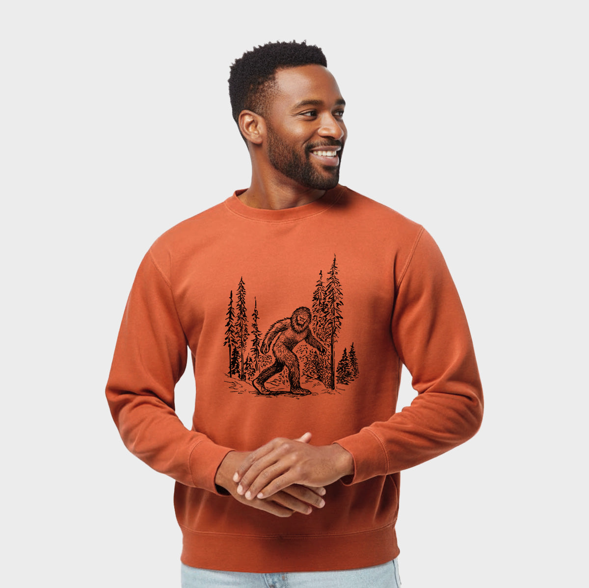 Model wearing an amber orange pigment dyed crewneck sweatshirt printed with a black ink illustration of Bigfoot walking through a pine forest.