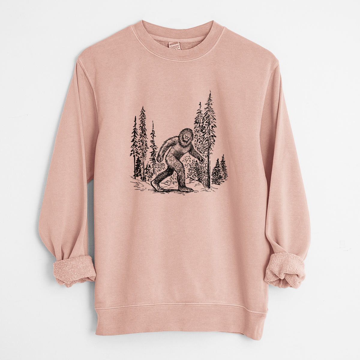 Dusty pink pigment dyed crewneck sweatshirt laid flat with the sleeves rolled, printed with a black ink illustration of Bigfoot walking through a pine forest.