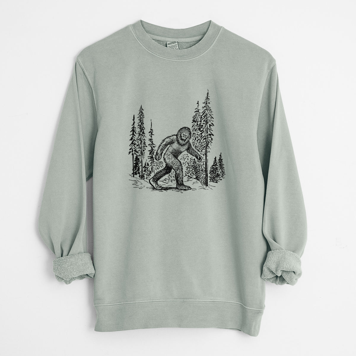 Sage green pigment dyed crewneck sweatshirt laid flat with the sleeves rolled, printed with a black ink illustration of Bigfoot walking through a pine forest.
