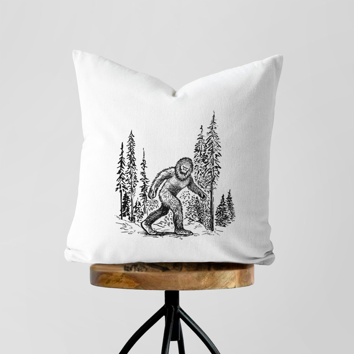 White pillow cover printed with a black ink illustration of Bigfoot walking through a pine forest, on a wooden stool.