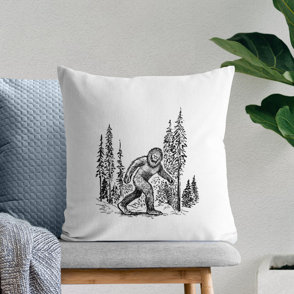 White pillow cover printed with a black ink illustration of Bigfoot walking through a pine forest, on a bench beside plants.