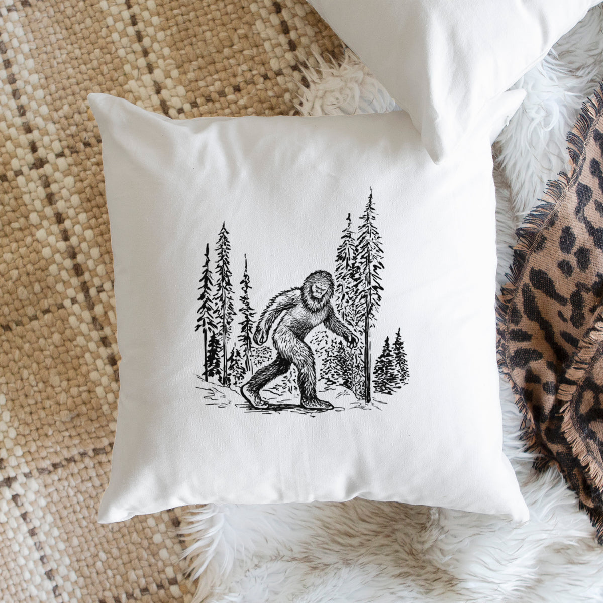 White pillow cover printed with a black ink illustration of Bigfoot walking through a pine forest, on a woven chair.