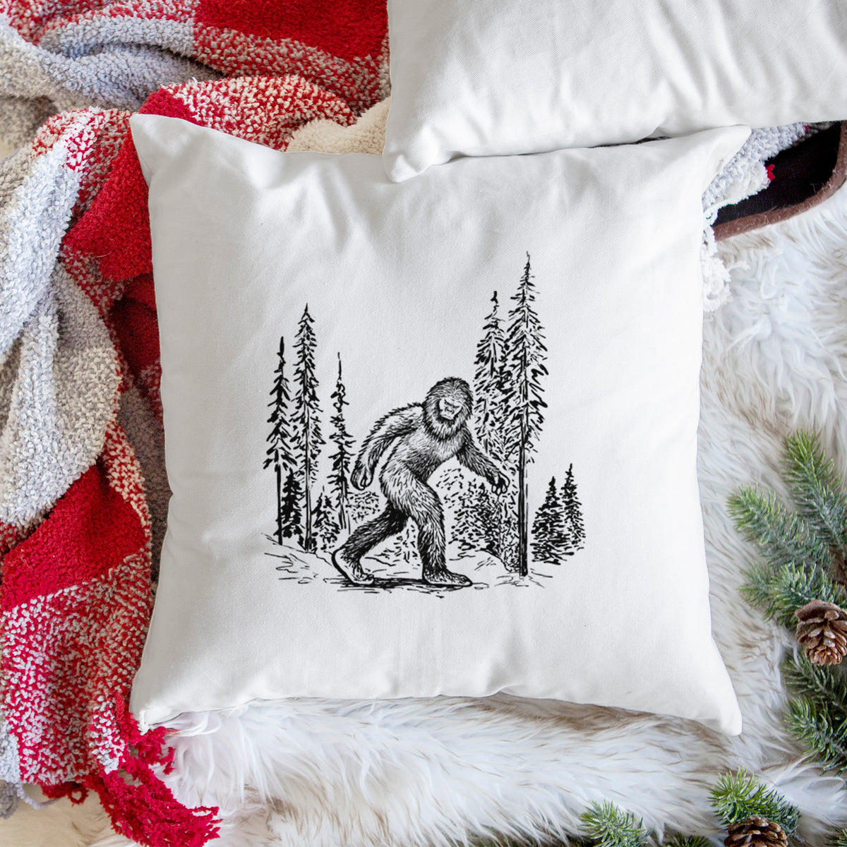 White pillow cover printed with a black ink illustration of Bigfoot walking through a pine forest, on a woven chair.