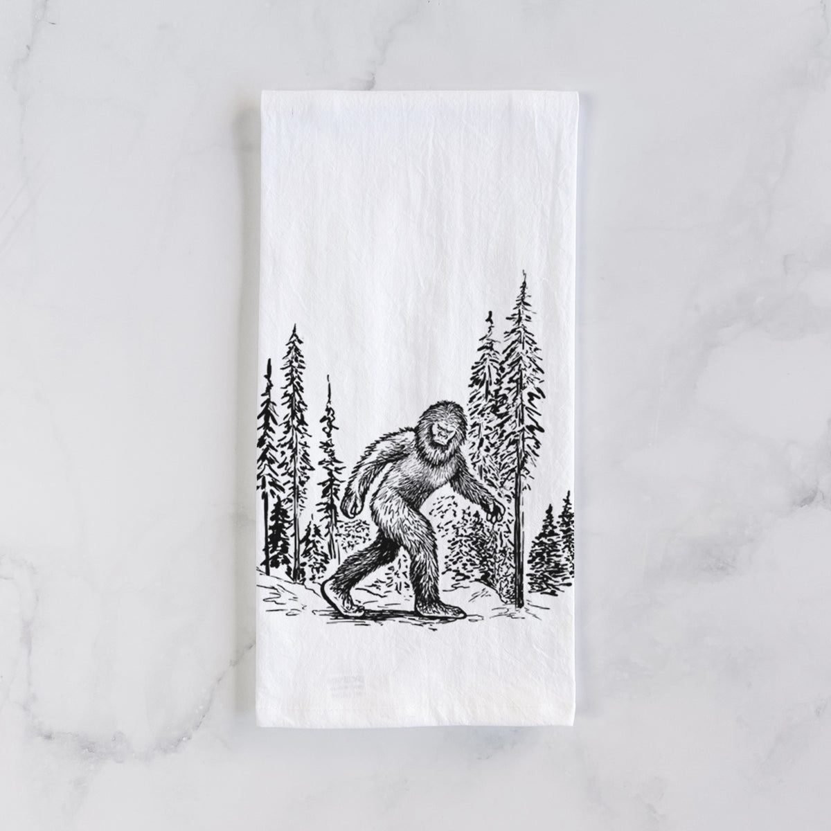 White cotton tea towel laid flat on marble, printed with a black ink illustration of Bigfoot walking through a pine forest.
