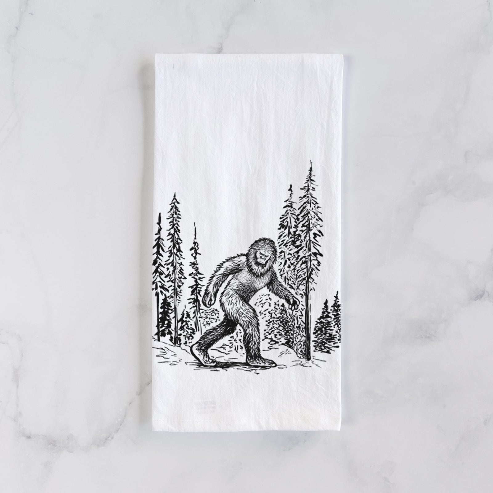 White cotton tea towel printed with a black ink illustration of Bigfoot walking through a pine forest, on a marble counter beside a round wooden cutting board and greenery.