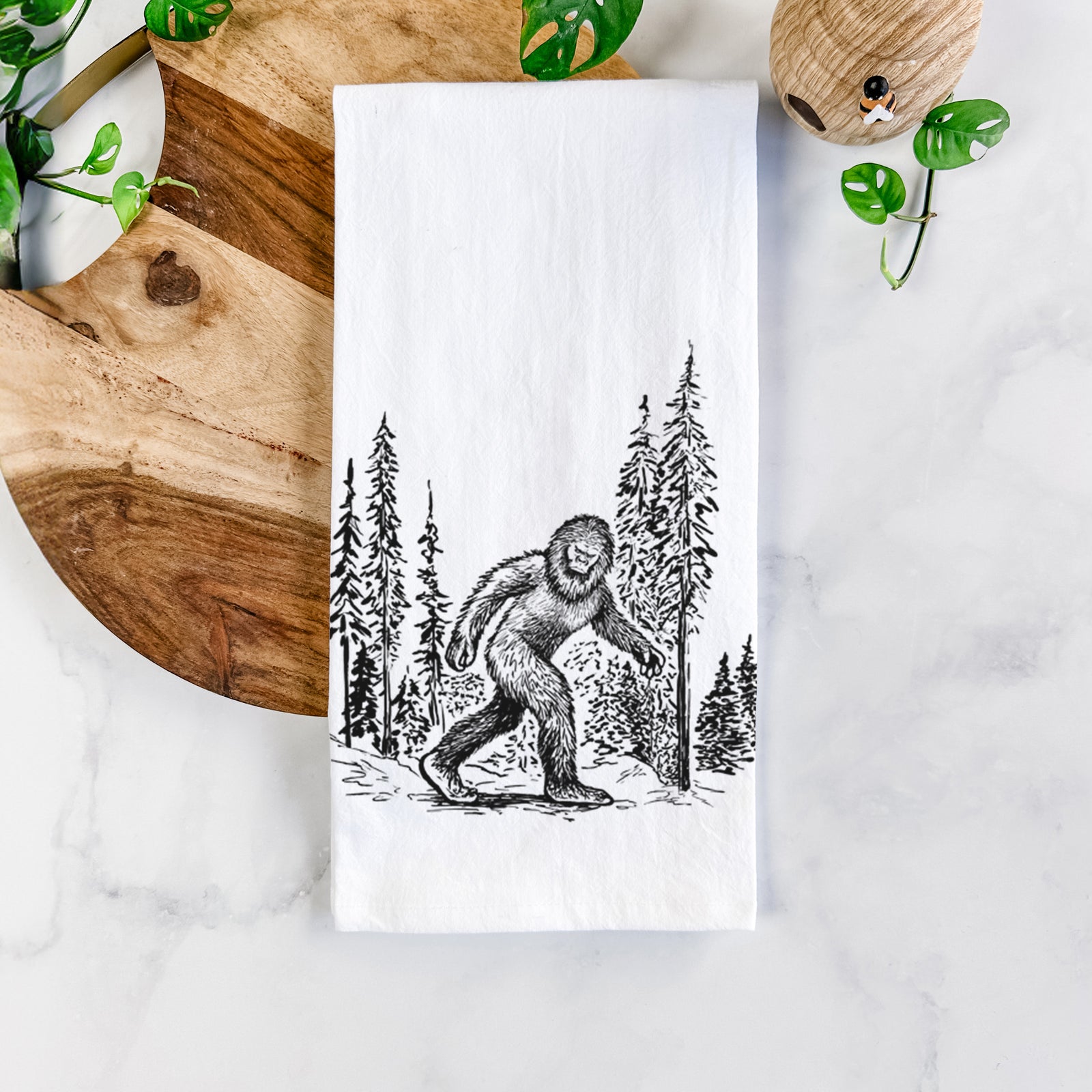 White cotton tea towel printed with a black ink illustration of Bigfoot walking through a pine forest, on a marble counter beside a round wooden cutting board and greenery.