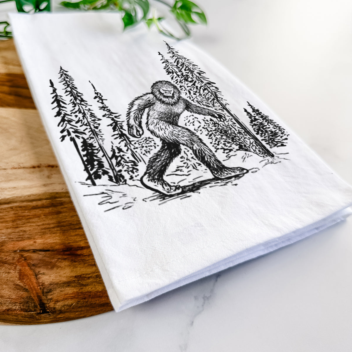Close-up of a folded white tea towel on a wooden board, showing a black ink illustration of Bigfoot walking through a pine forest.
