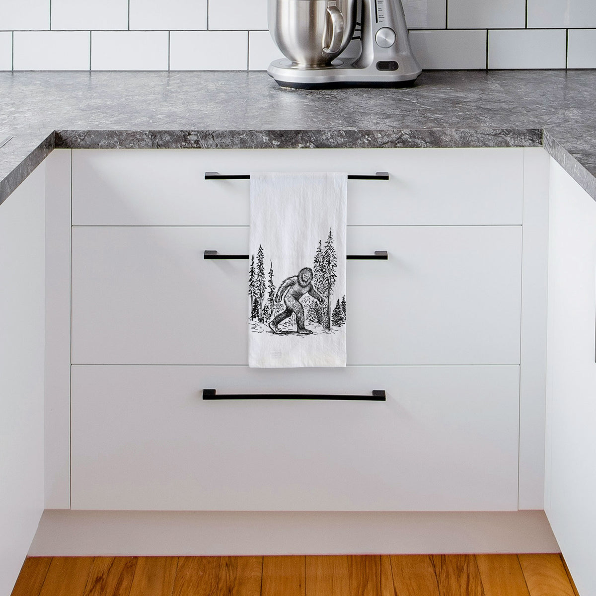 White tea towel printed with a black ink illustration of Bigfoot walking through a pine forest, hanging from a drawer handle in a bright white kitchen.