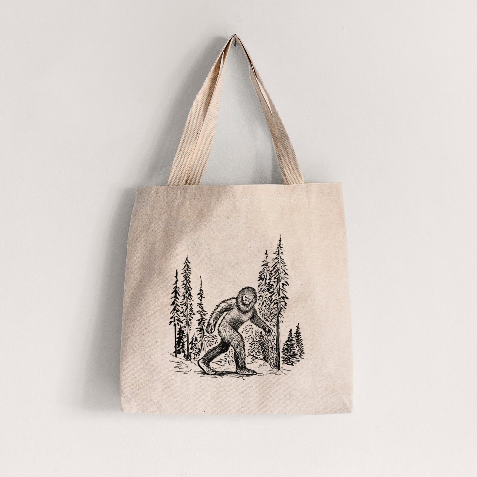 Natural canvas tote bag printed with a black ink illustration of Bigfoot walking through a pine forest.