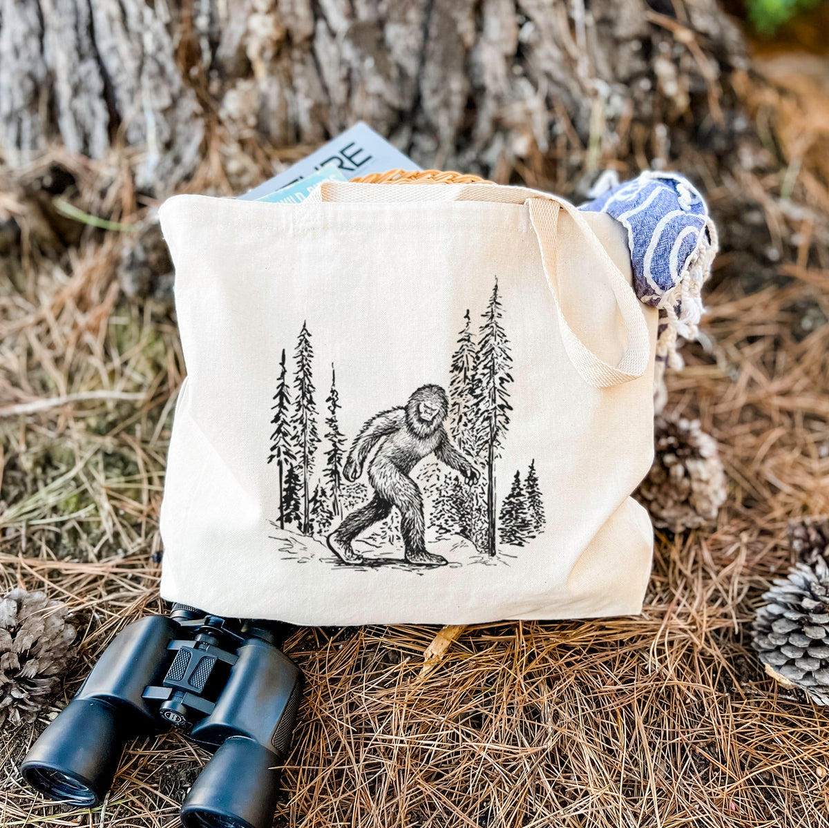 Canvas tote bag printed with a black ink illustration of Bigfoot walking through a pine forest, resting on dry grass beside a pair of binoculars.
