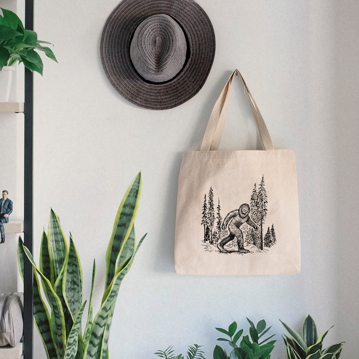 Canvas tote bag printed with a black ink illustration of Bigfoot walking through a pine forest, hanging on a wall hook below a hat, next to potted plants.