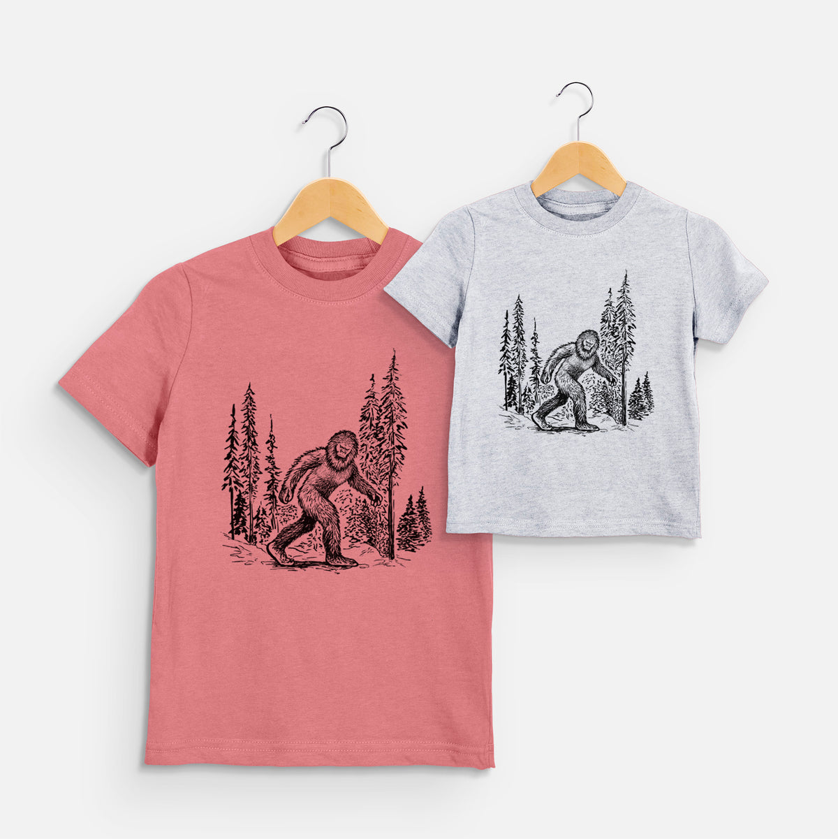 Two youth t-shirts in different colors on hangers, printed with a black ink illustration of Bigfoot walking through a pine forest.
