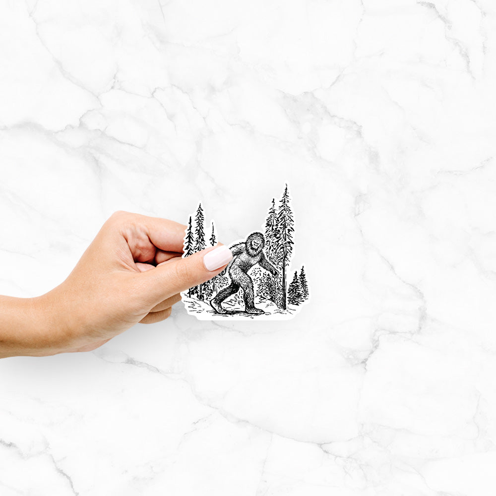 Hand holding a 4 inch vinyl decal of a black ink illustration of Bigfoot walking through a pine forest.