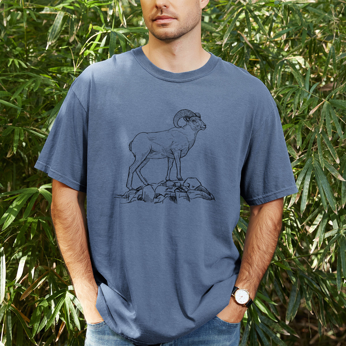 Man wearing a Comfort Colors t-shirt printed with a black ink illustration of a bighorn ram with curled horns standing on rocks, outdoors.