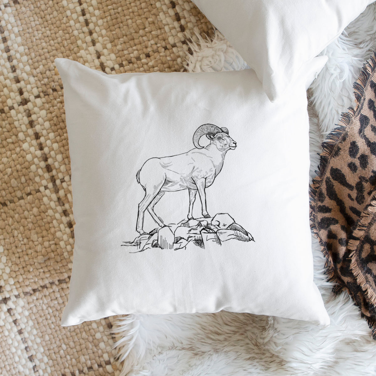 White pillow cover printed with a black ink illustration of a bighorn ram with curled horns standing on rocks, on a woven chair.