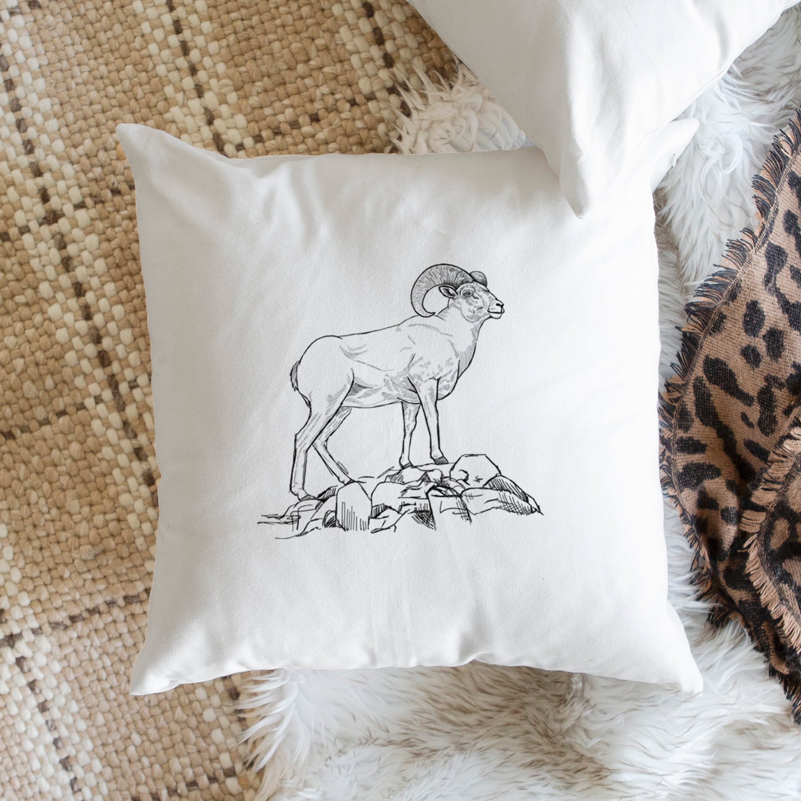 White pillow cover printed with a black ink illustration of a bighorn ram with curled horns standing on rocks, on a bench beside plants.
