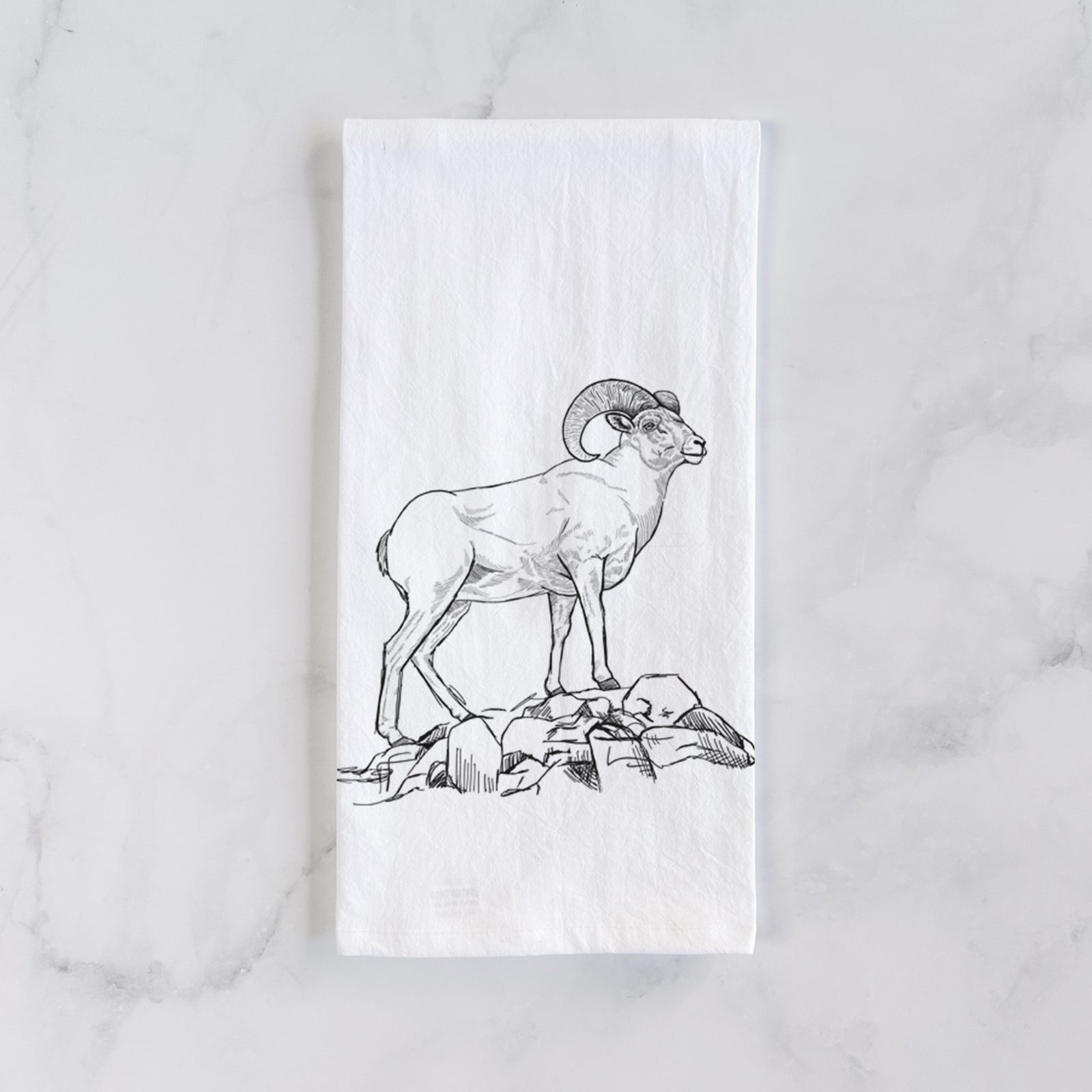 White cotton tea towel printed with a black ink illustration of a bighorn ram with curled horns standing on rocks, on a marble counter beside a round wooden cutting board and greenery.