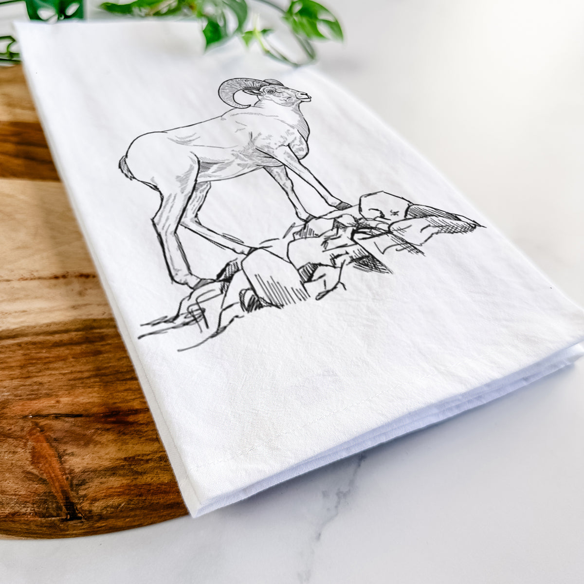 Close-up of a folded white tea towel on a wooden board, showing a black ink illustration of a bighorn ram with curled horns standing on rocks.