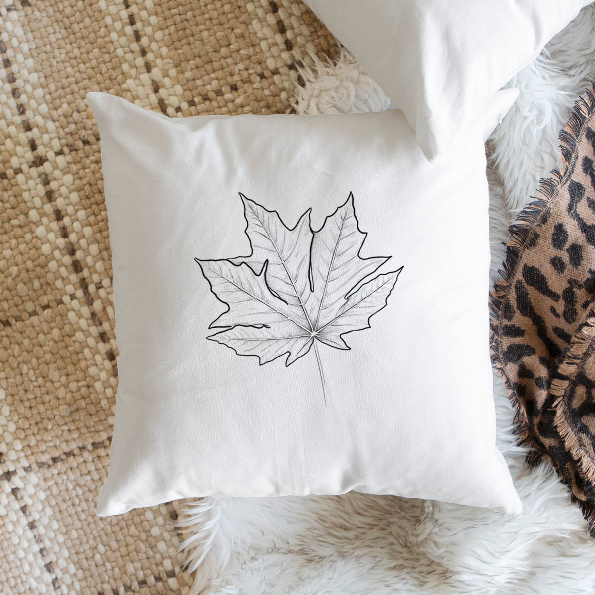 White pillow cover printed with a black ink line drawing of a bigleaf maple leaf, on a woven chair.