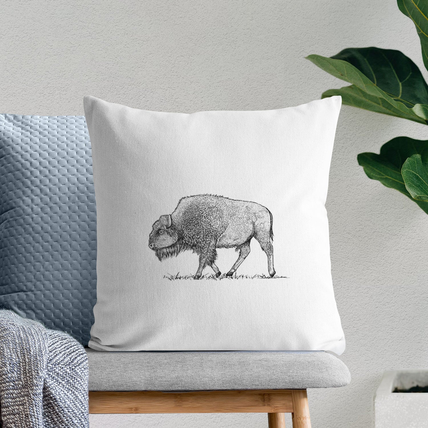 White pillow cover printed with a detailed black ink illustration of an American bison walking through grass, on a bench beside plants.