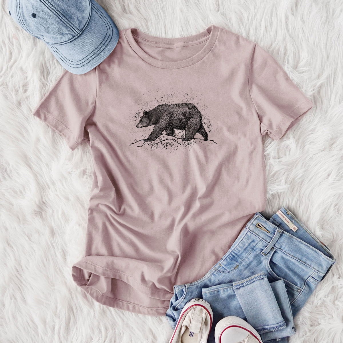 Women's relaxed fit cotton t-shirt printed with a detailed black ink sketch of a black bear walking across rocky ground, styled flat on a white faux fur rug with jeans.