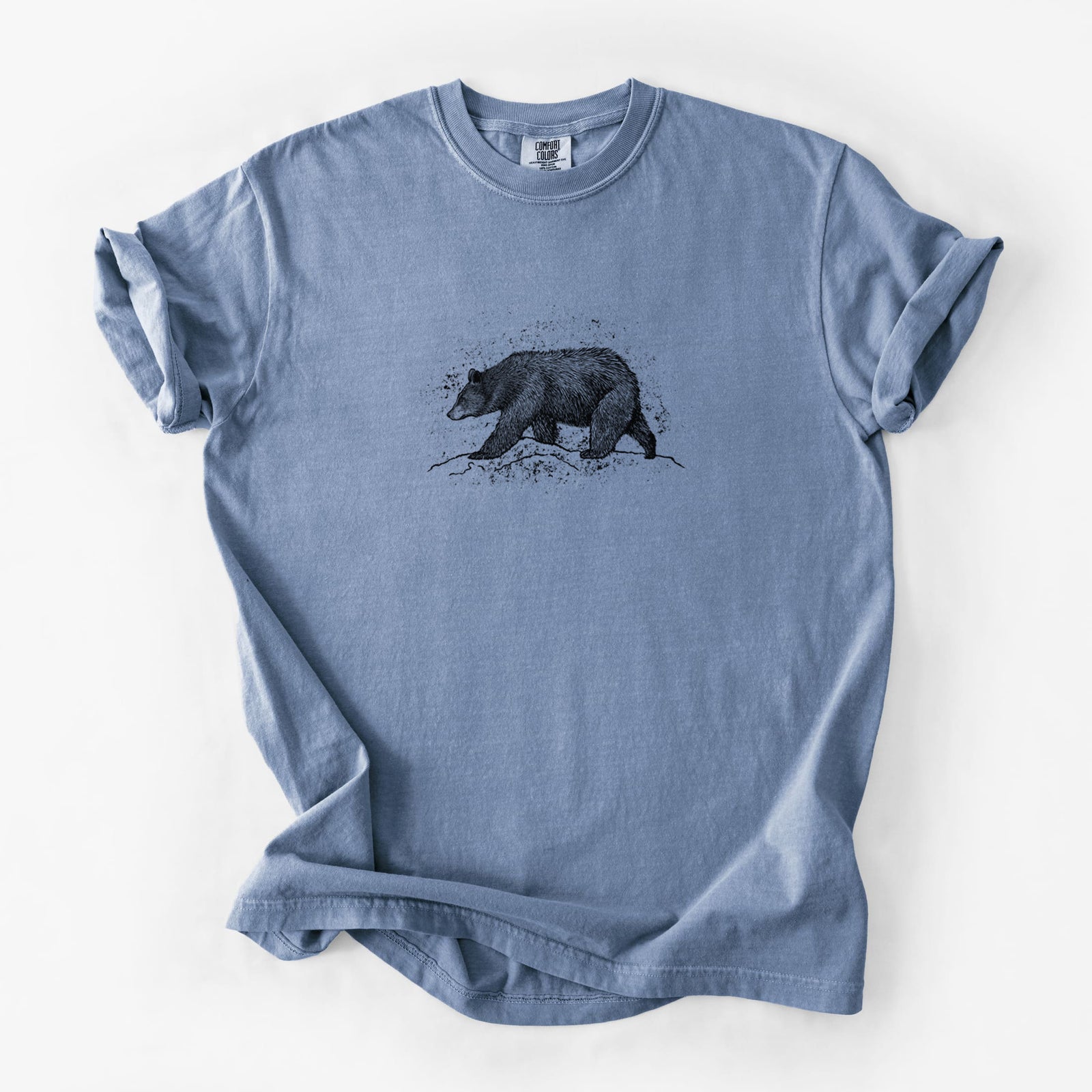 Blue jean Comfort Colors t-shirt laid flat, printed with a detailed black ink sketch of a black bear walking across rocky ground.