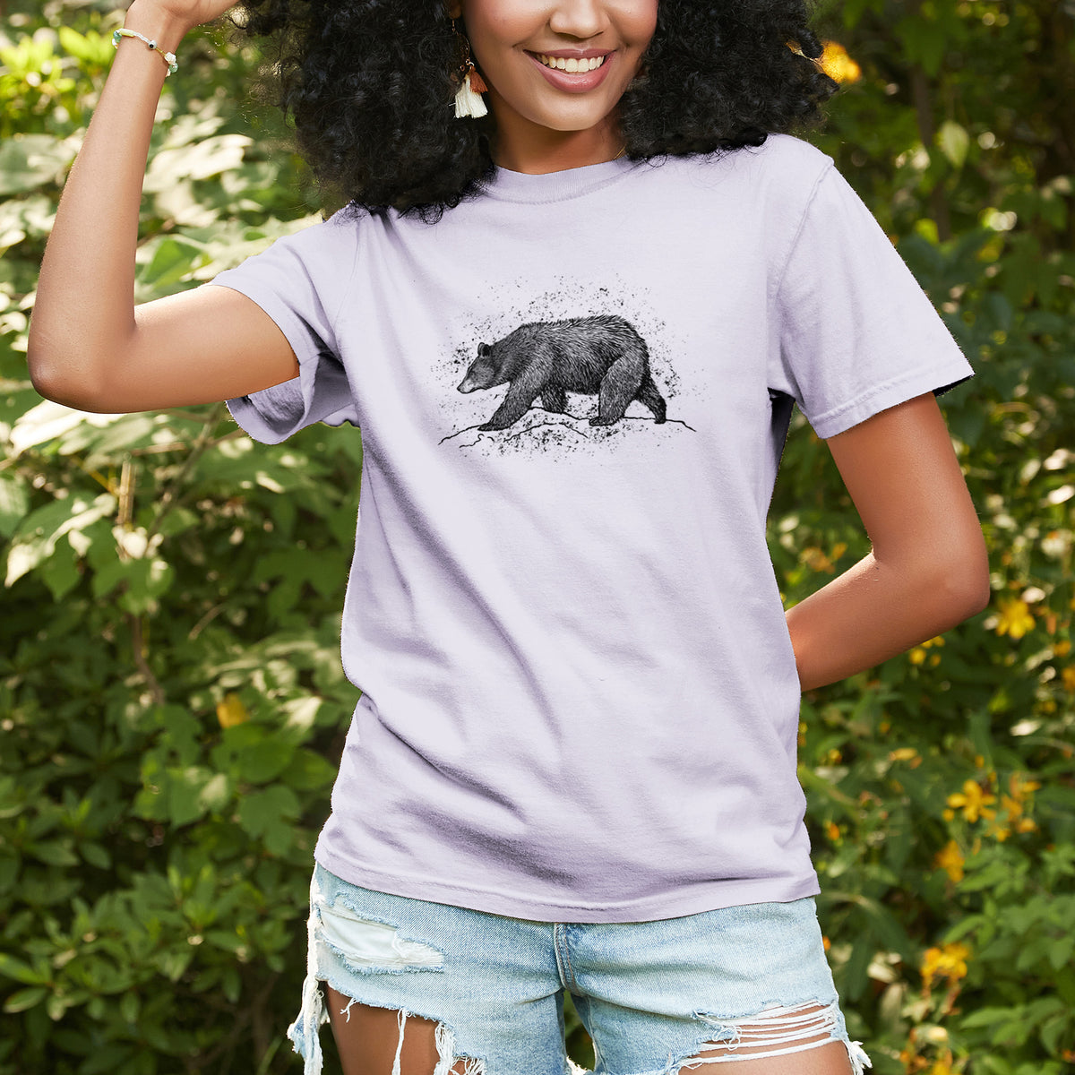 Woman wearing a Comfort Colors t-shirt printed with a detailed black ink sketch of a black bear walking across rocky ground, outdoors.