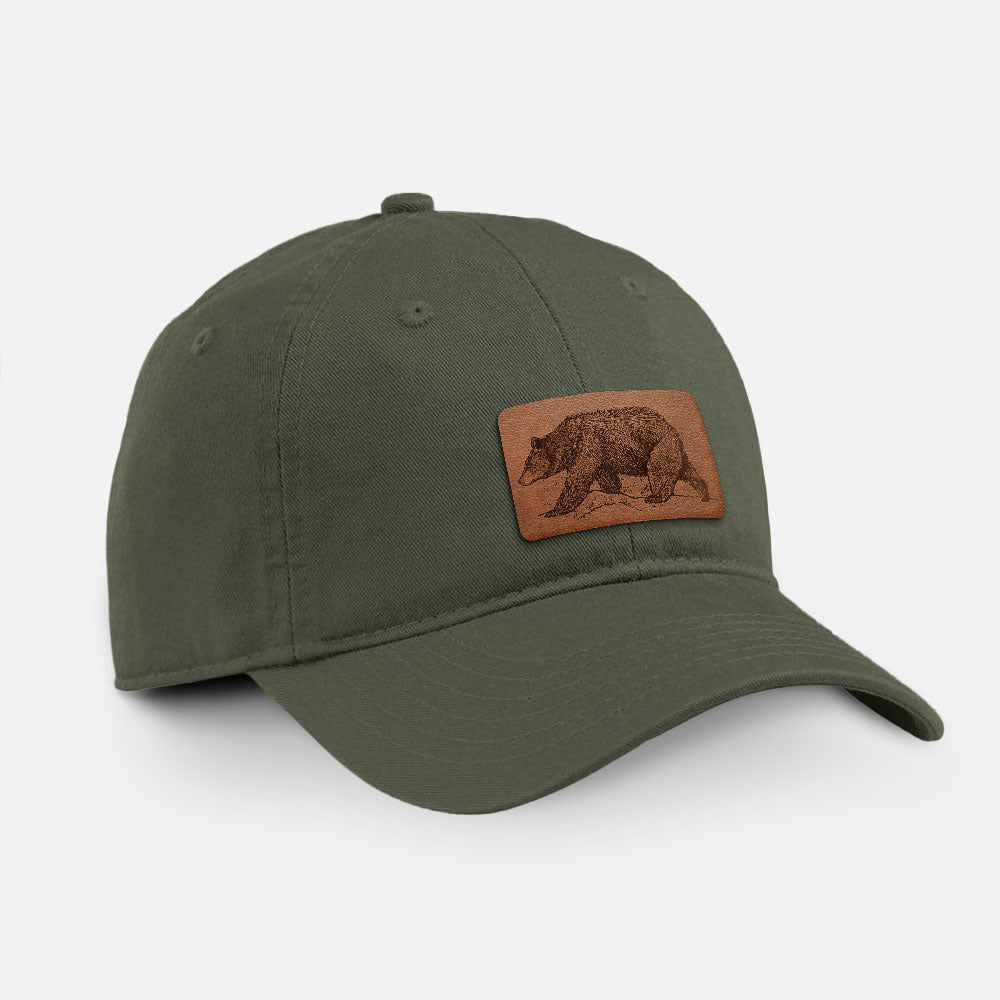 Olive cotton baseball hat with a detailed black ink sketch of a black bear walking across rocky ground on the front.