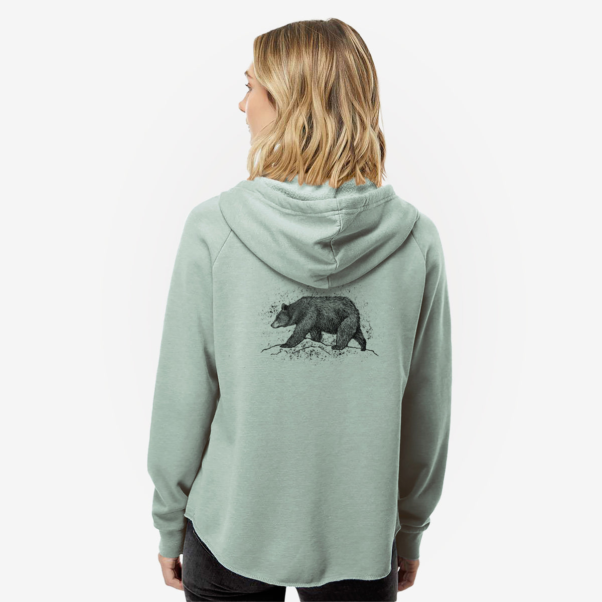 Woman wearing a sage green women's Cali Wave zip-up sweatshirt printed with a detailed black ink sketch of a black bear walking across rocky ground, seen from behind.