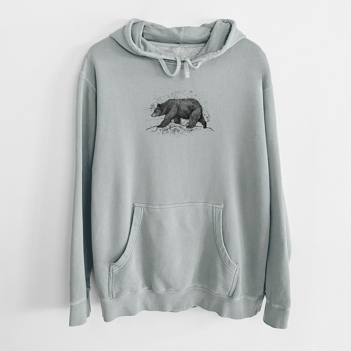 Sage green pigment dyed hoodie laid flat, printed with a detailed black ink sketch of a black bear walking across rocky ground.