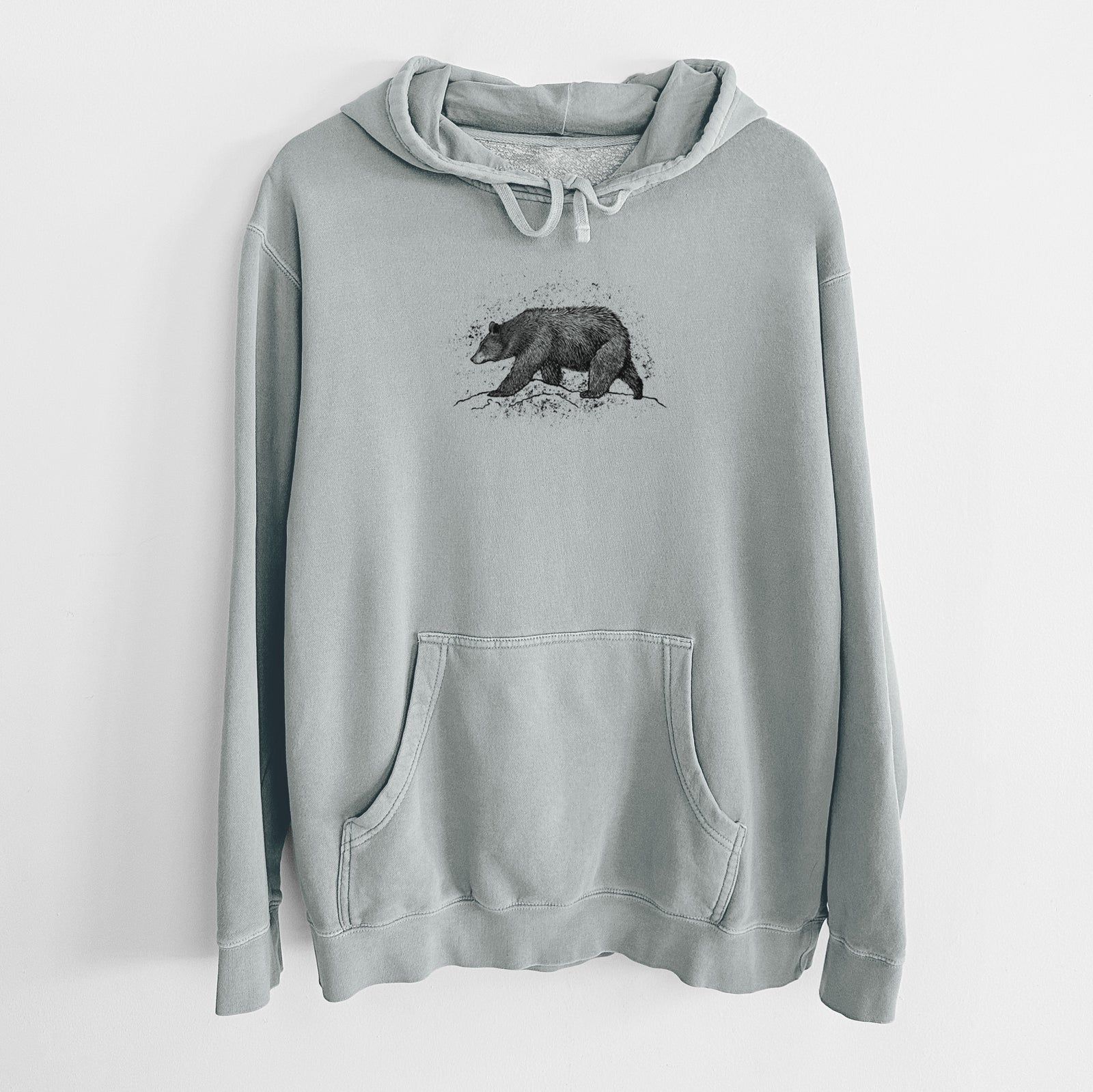 Sage green pigment dyed hoodie laid flat, printed with a detailed black ink sketch of a black bear walking across rocky ground.