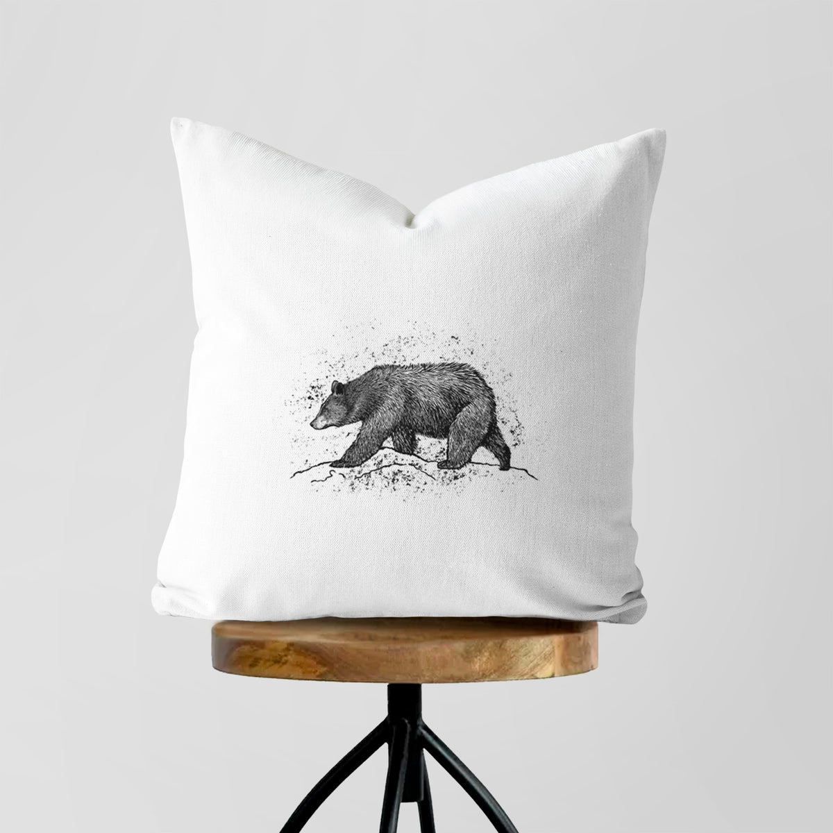White pillow cover printed with a detailed black ink sketch of a black bear walking across rocky ground, on a wooden stool.