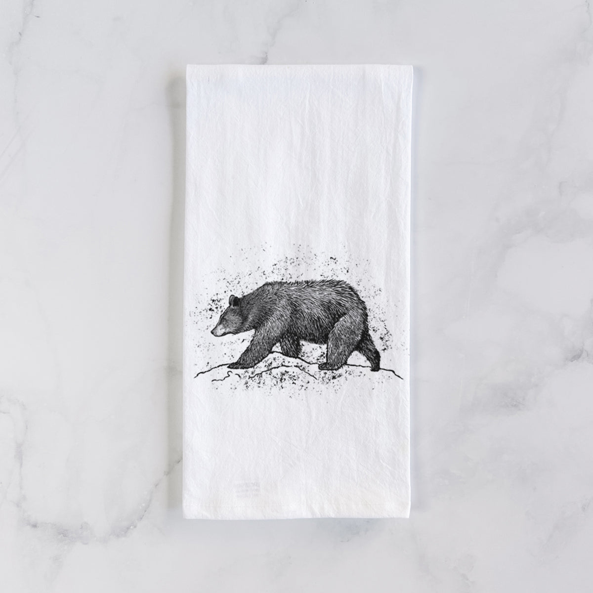 White cotton tea towel laid flat on marble, printed with a detailed black ink sketch of a black bear walking across rocky ground.