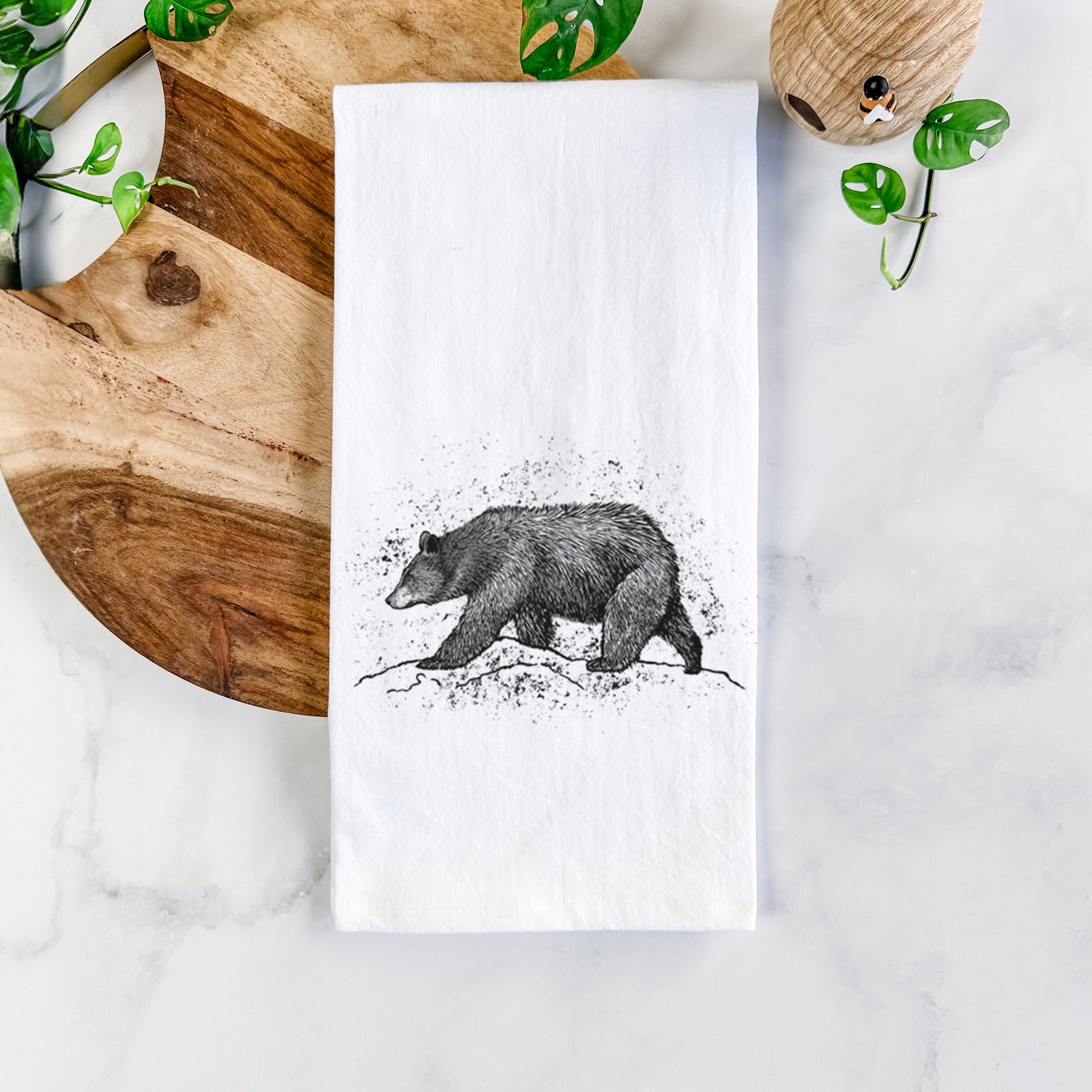 White cotton tea towel printed with a detailed black ink sketch of a black bear walking across rocky ground, on a marble counter beside a round wooden cutting board and greenery.
